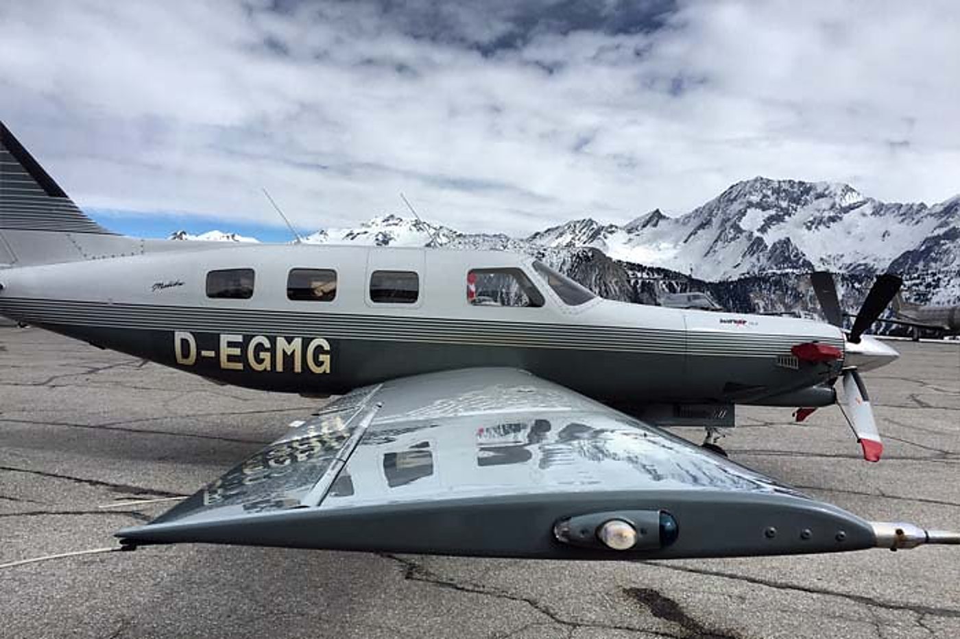 Un petit avion à hélice gris stationné sur le tarmac d'un aérodrome de montagne, avec des sommets enneigés et un ciel nuageux en arrière-plan.