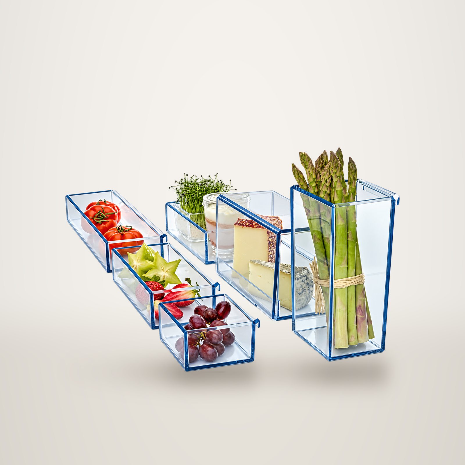 Plusieurs bacs en verre transparent aux bords bleutés contenant des tomates, des fruits, du raisin, des fromages, des pousses et des asperges sur fond blanc.