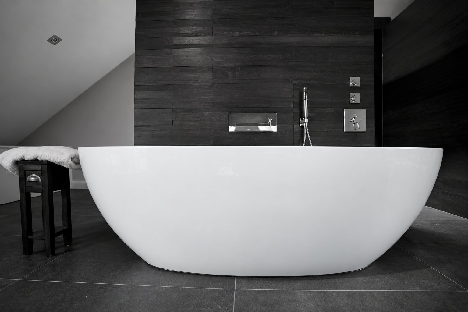 An oval white freestanding bath on dark tiles in front of a dark wooden wall with wall-mounted taps, beside a small stool holding a towel.