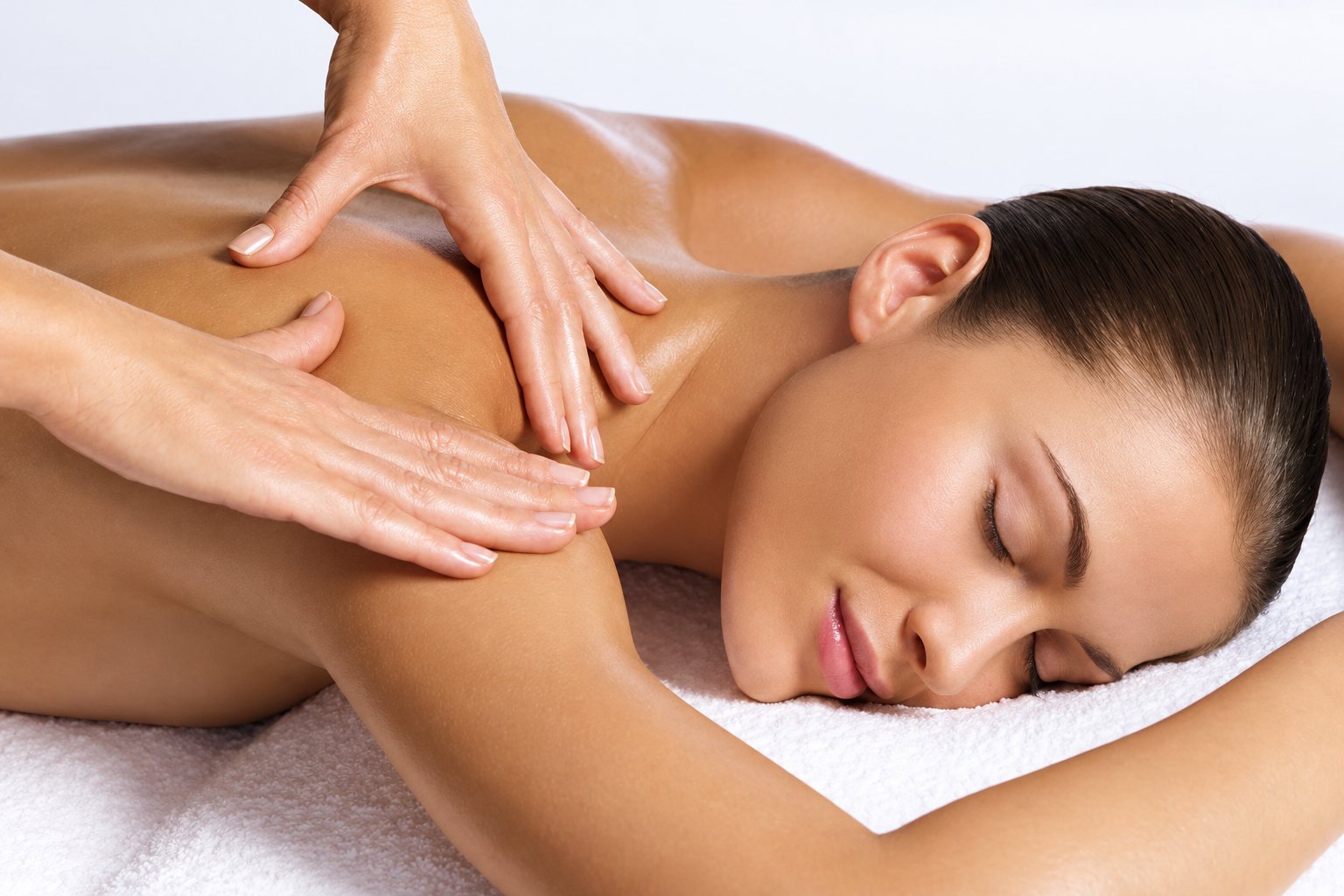 A woman lying on a white towel with her eyes closed receives a relaxing back and shoulder massage.