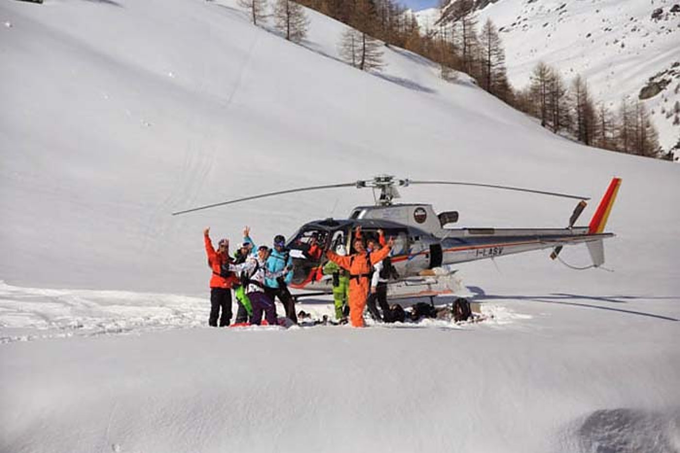 Un groupe de skieurs en tenues colorées lève les bras devant un hélicoptère posé dans une vallée enneigée.