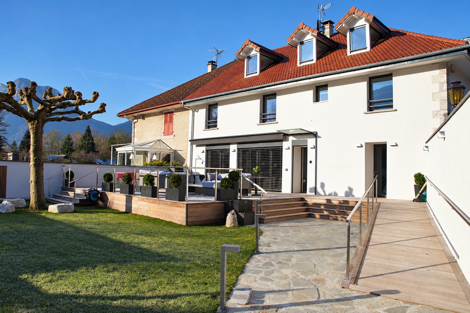 Une grande maison blanche à toit de tuiles rouges avec lucarnes, une terrasse en bois, une rampe d'accès et un jardin engazonné devant des montagnes.