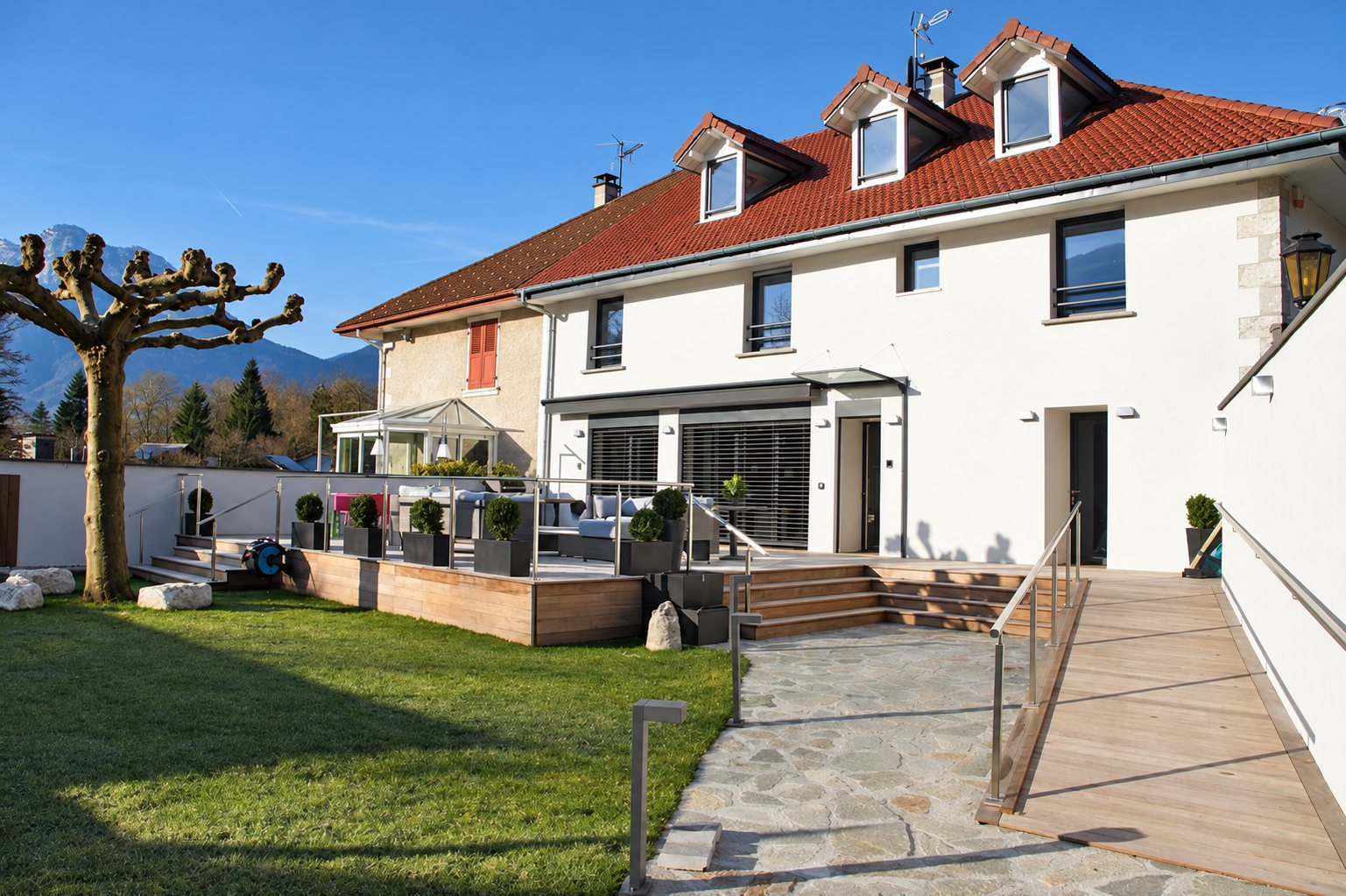 White façade of a renovated house with a red tile roof, a wooden terrace, an access ramp, a garden and mountains in the distance.