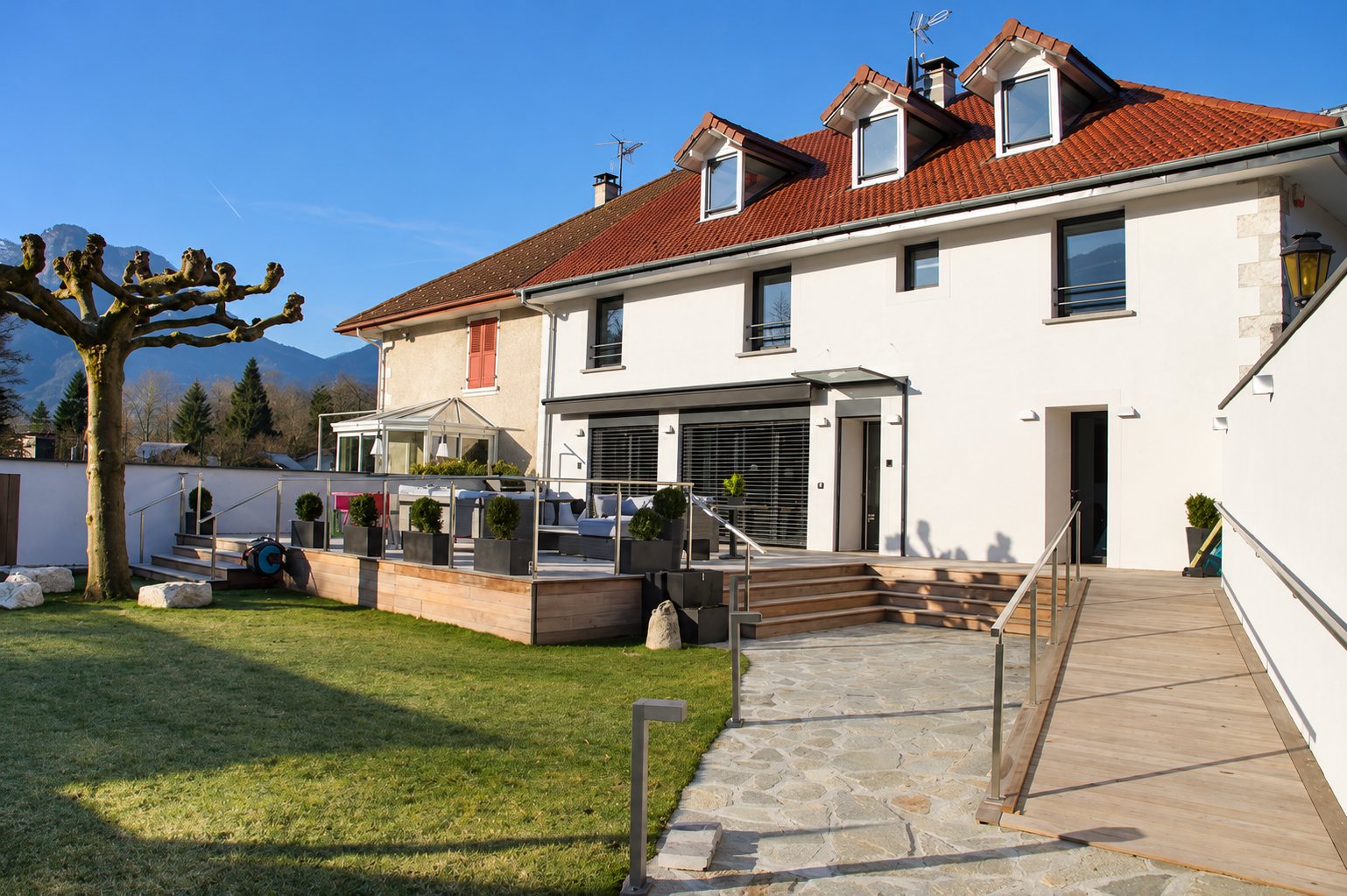 White façade of a house with a red tile roof, a wooden terrace, an access ramp, a lawn and a pruned plane tree, in front of mountains.