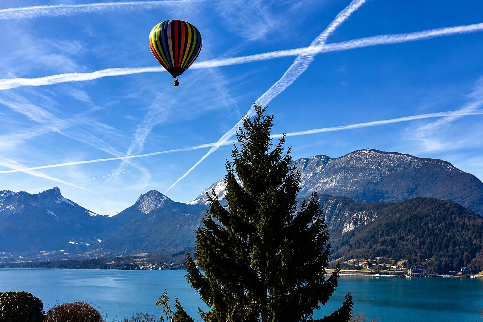 A hot-air balloon with coloured bands floating in a blue sky streaked with white trails above a lake and mountains, with a tall fir tree in the foreground.