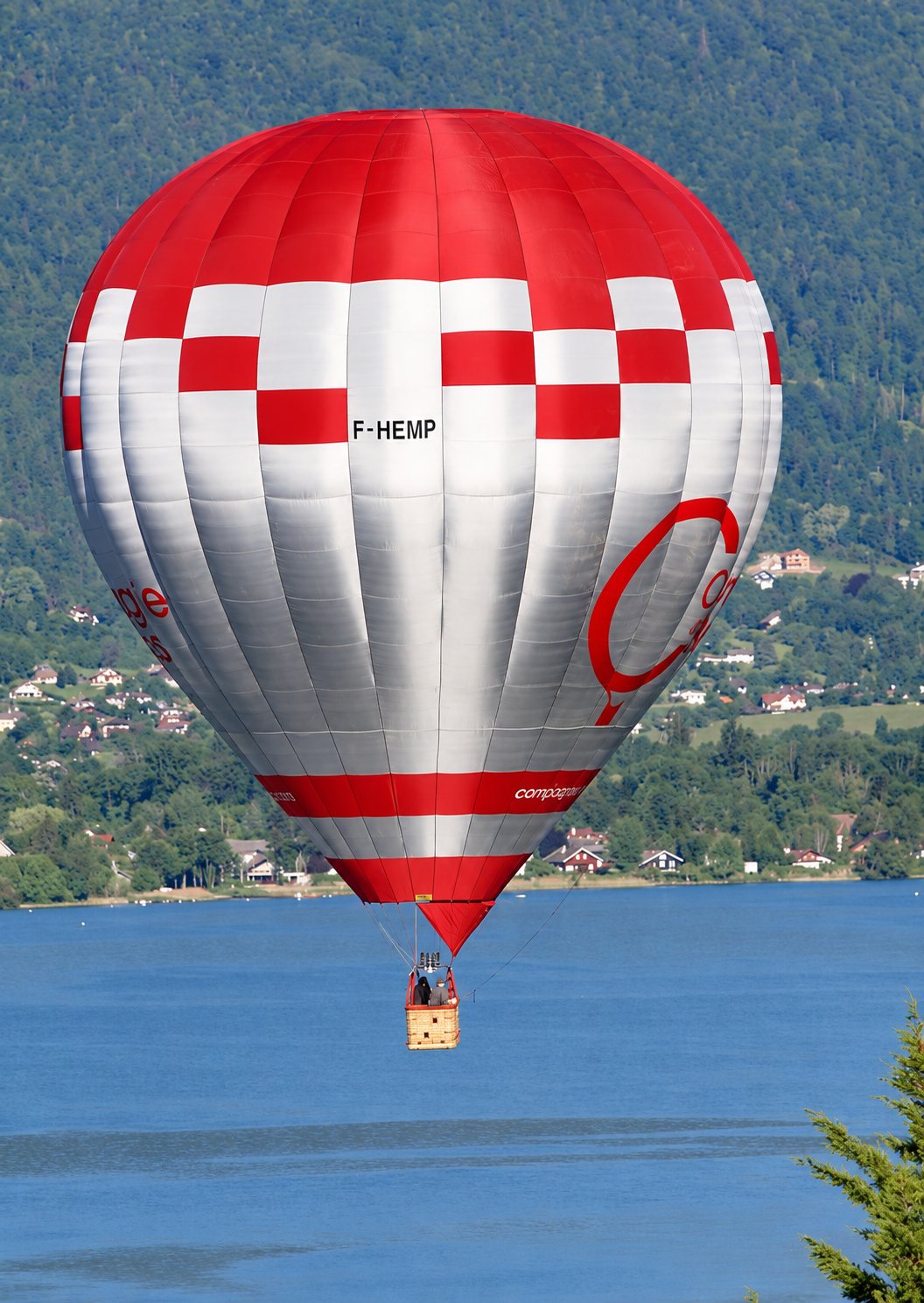 Une montgolfière à damier rouge, blanc et argent, avec sa nacelle en osier, vole au-dessus d'un lac bleu devant des collines boisées.