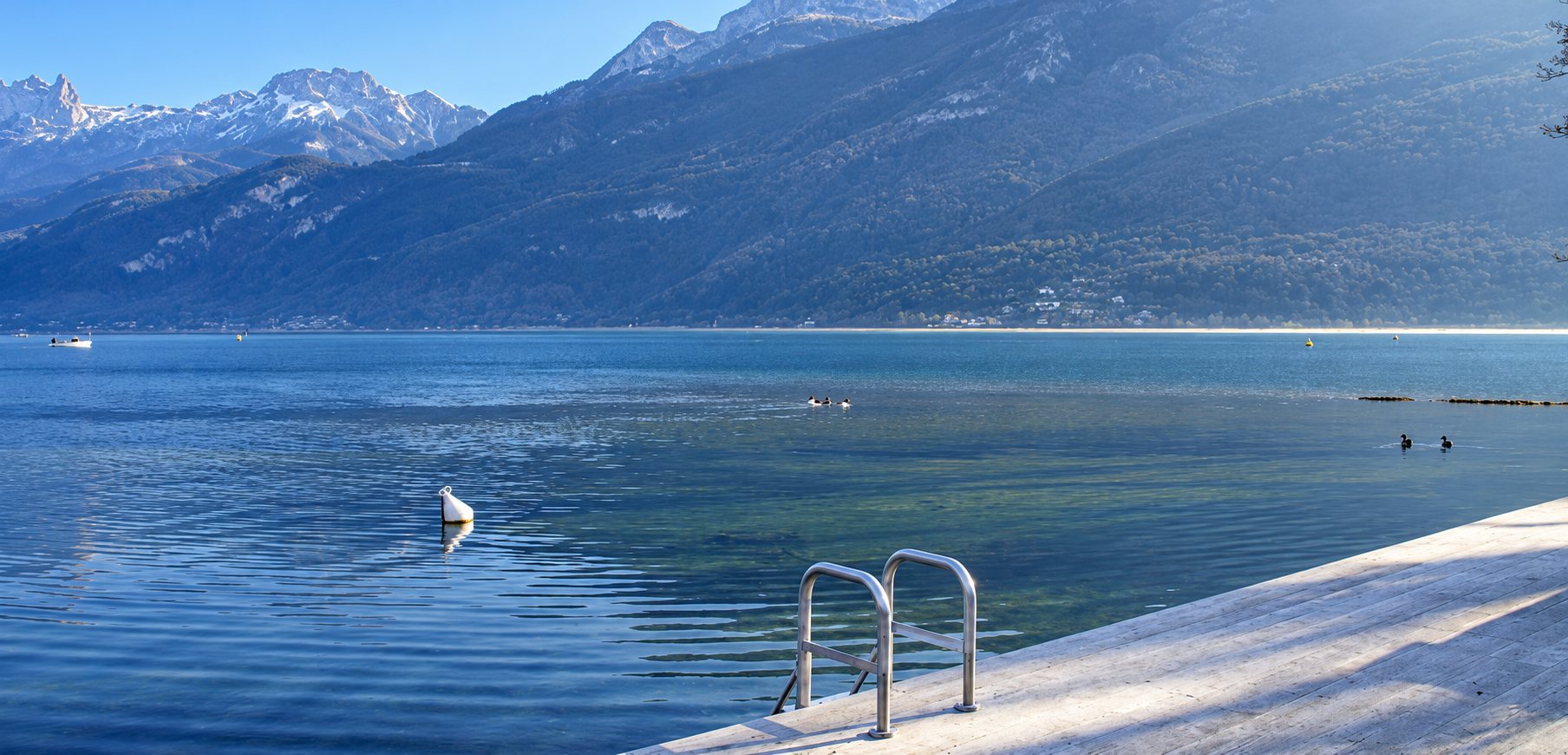 Un ponton en bois clair équipé d'une échelle en inox s'avance sur un lac calme entouré de montagnes dans la brume.