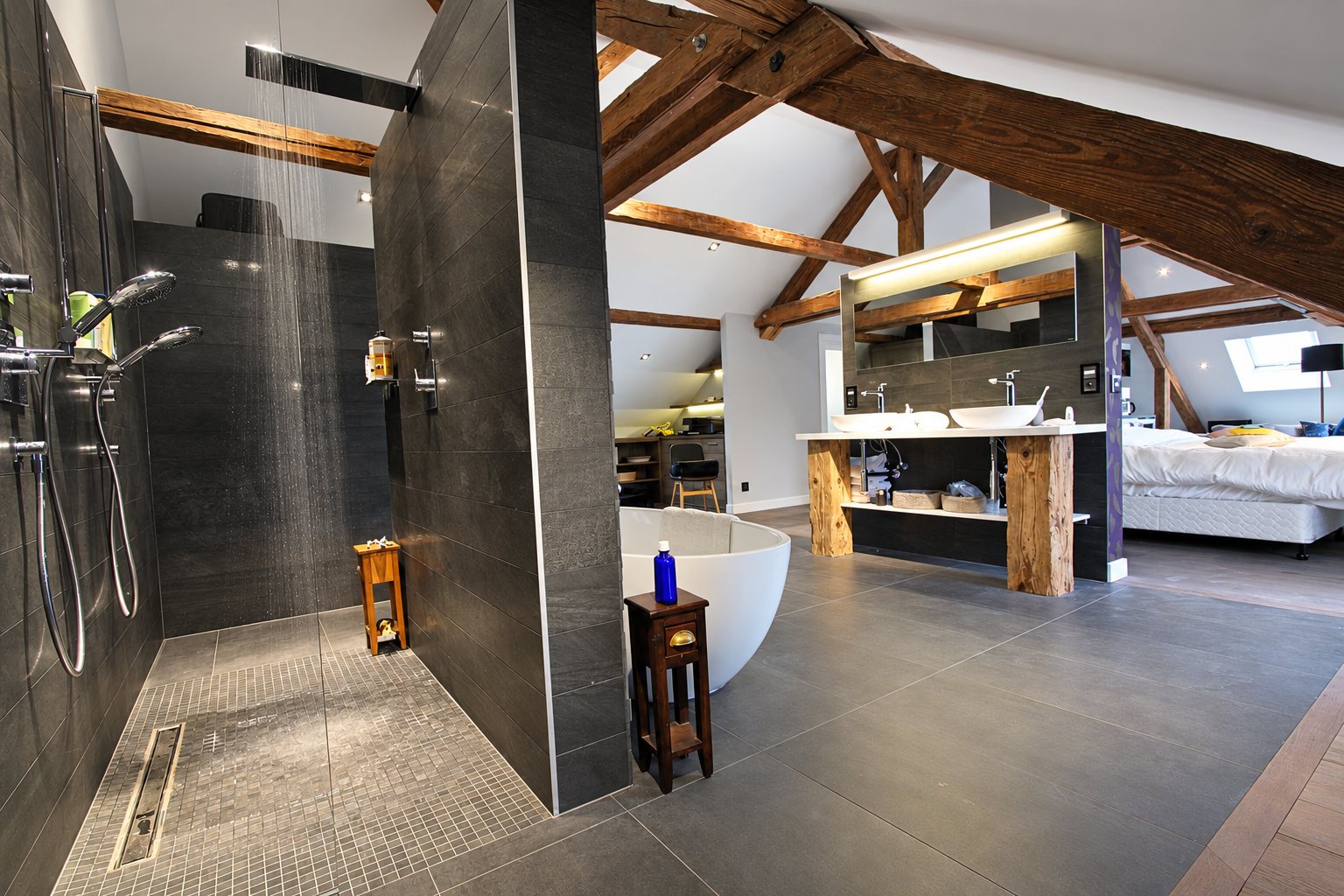Salle de bain moderne sous charpente apparente, avec douche à l'italienne, baignoire ovale et double vasque ouverte sur la chambre.