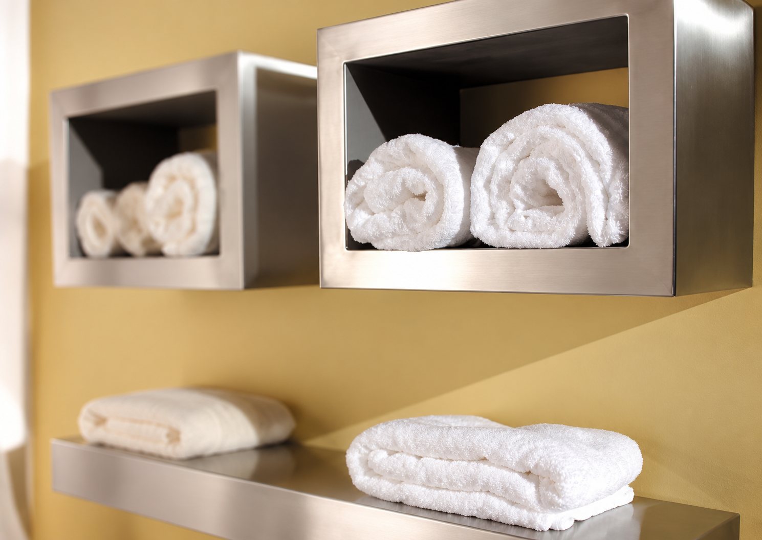 Soft white towels rolled in brushed steel wall niches on a yellow wall, with a folded towel on a shelf below.