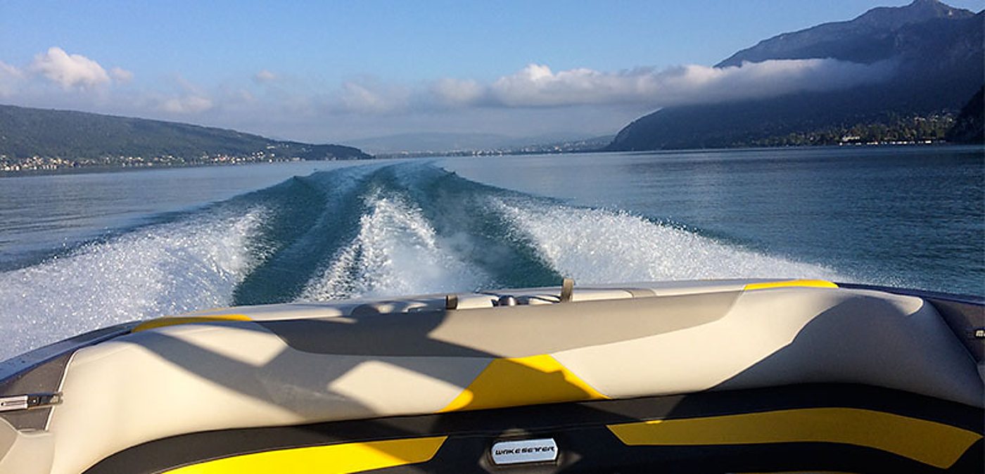 Vue depuis l'arrière d'un bateau à moteur gris et jaune, laissant un large sillage d'écume sur un lac bordé de montagnes.
