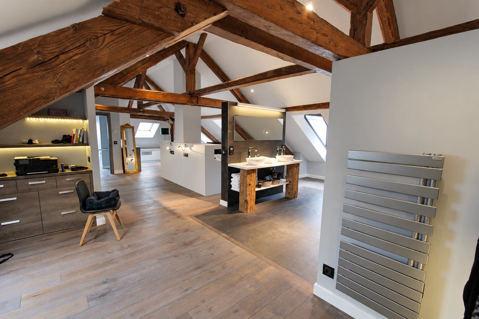 Vaste espace sous les combles avec poutres apparentes, parquet gris, double vasque posée sur un meuble en bois et radiateur sèche-serviettes en inox.