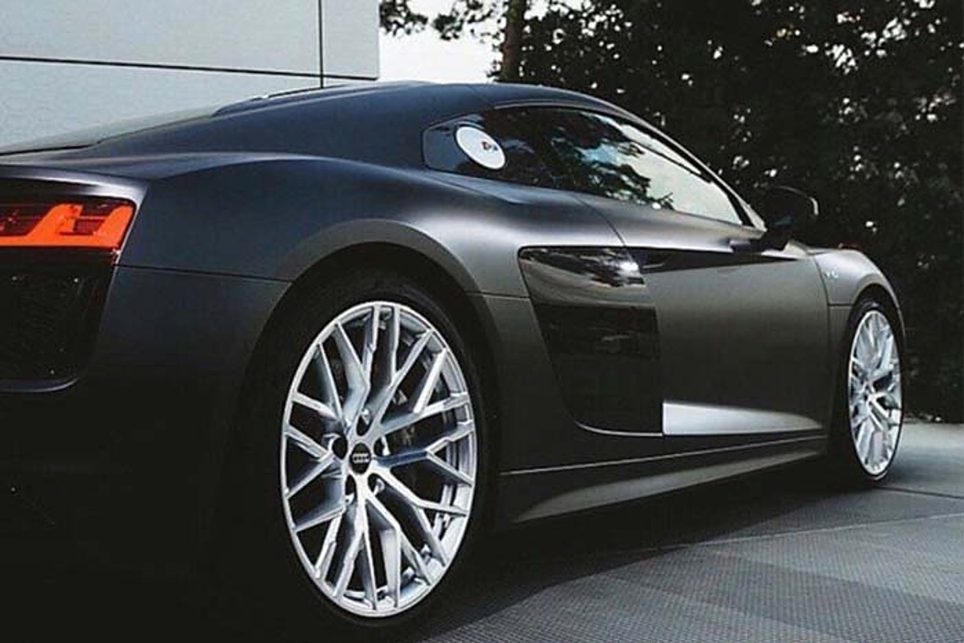 A matt grey sports car parked in front of a pale wall, seen from the rear three-quarter, with large silver fine-spoked wheels.