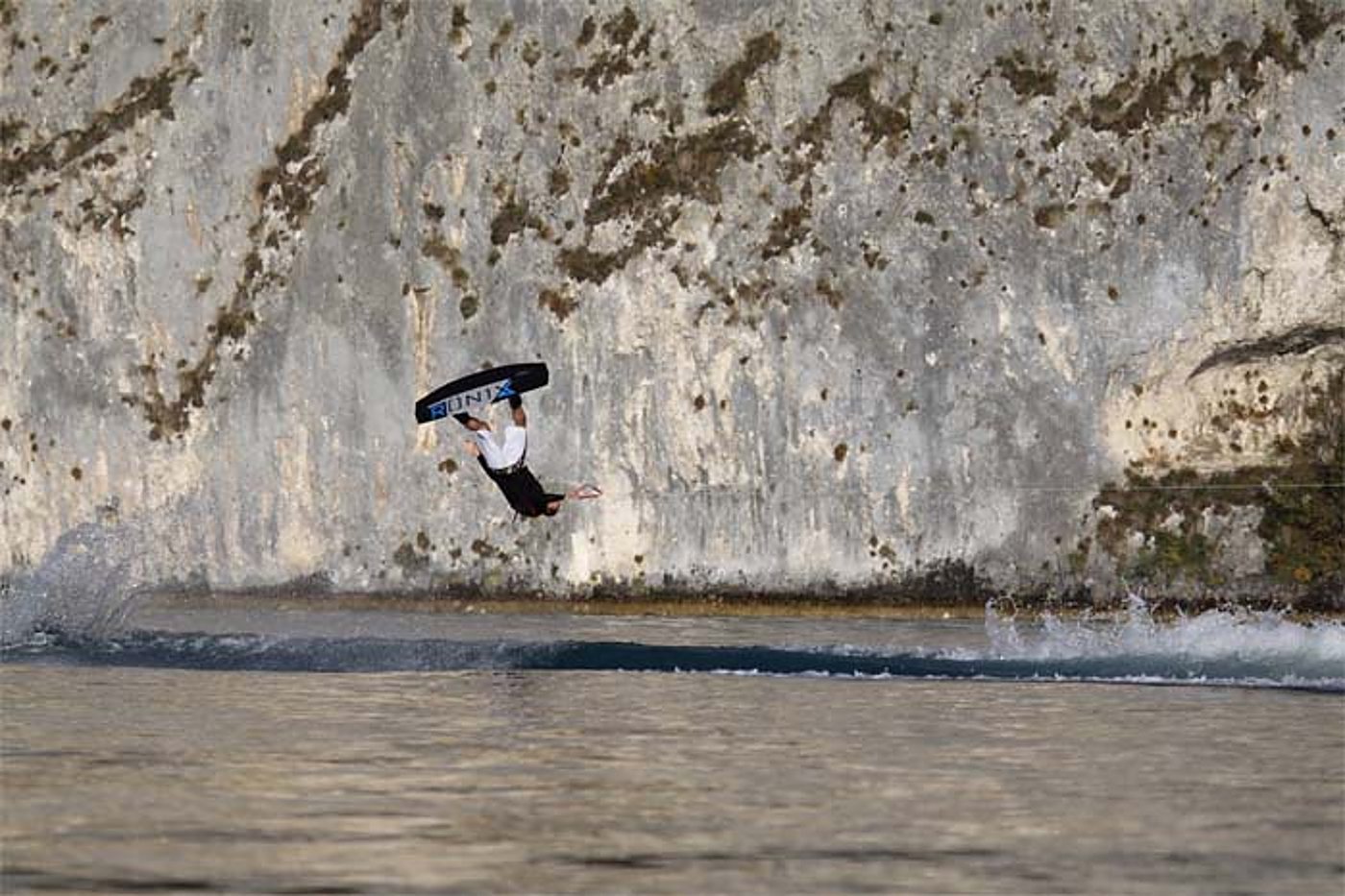 Un wakeboardeur est en l'air, la planche au-dessus de lui, en plein saut au-dessus de l'eau au pied d'une grande falaise rocheuse.